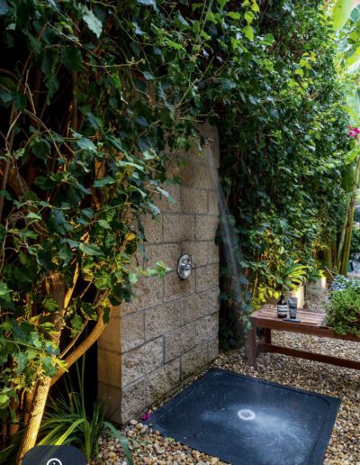 An outdoor shower with stone walls covered in climbing plants, featuring a wooden bench and a pebbled path.
